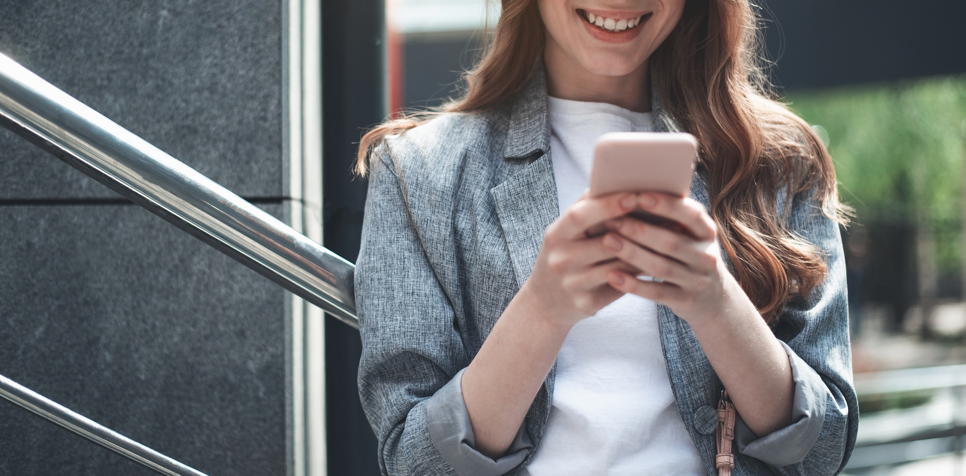 Waist up portrait of cheerful beautiful lady standing outside near the stairs. She is holding her smartphone and looking at it with joy